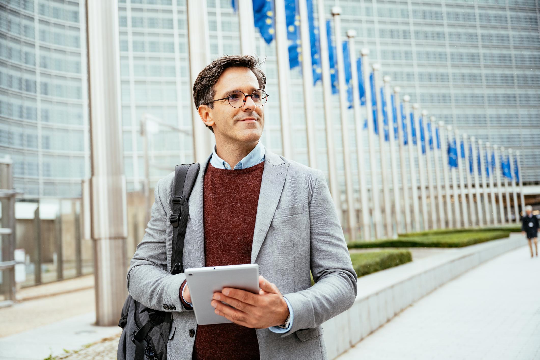 Mid adult white politician holding digital tablet near governmetnal building and using internet for business purposes. European union flags waving in the background